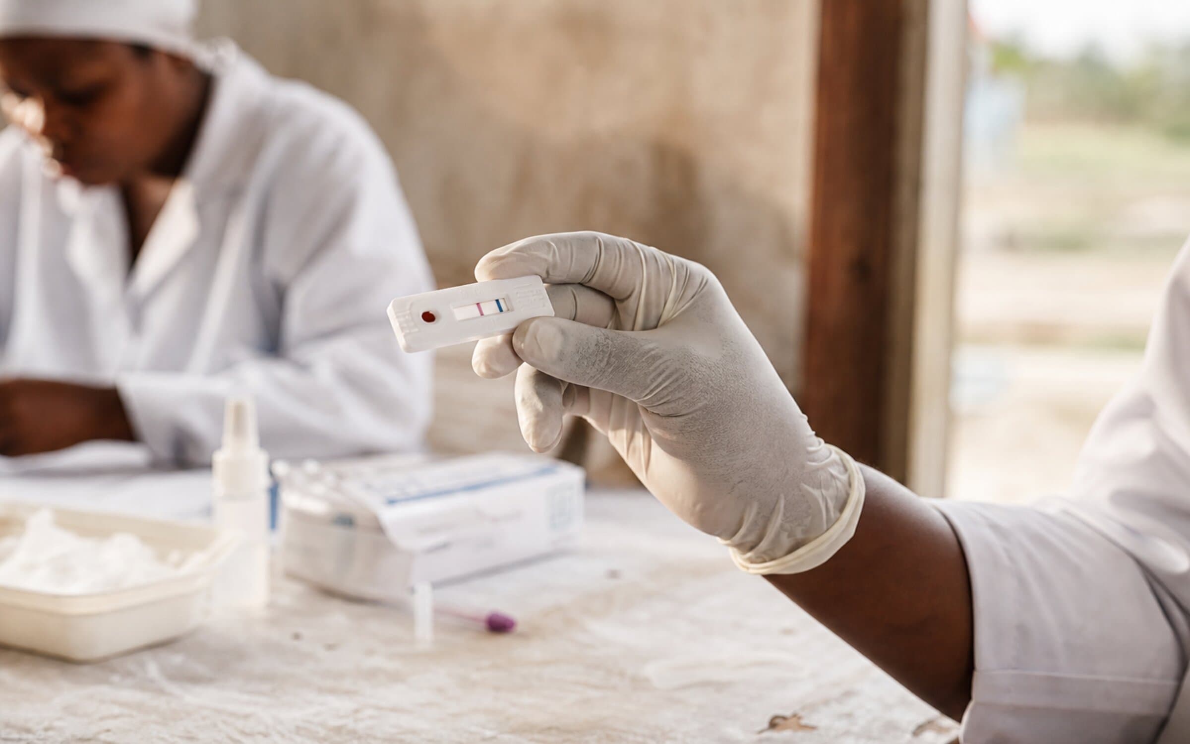 A gloved hand reading a rapid diagnostic test cassette in a community-clinic setting, showing the real-world conditions Audere's interpretation models are trained for.